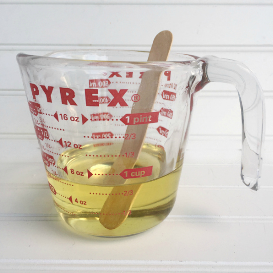 Melted soy wax in a glass measuring cup with a wooden craft stick used for stirring