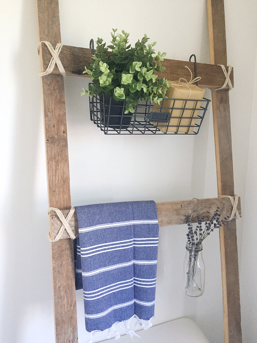 Rustic ladder displaying a wire basket with a plant, a Turkish towel, and a glass milk jar with dried lavender