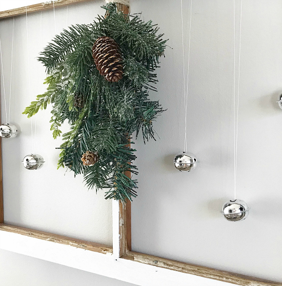 Old chippy white window hung on wall with pine greenery and jingle bells hanging from it. Perfect farmhouse wall decor for Christmas