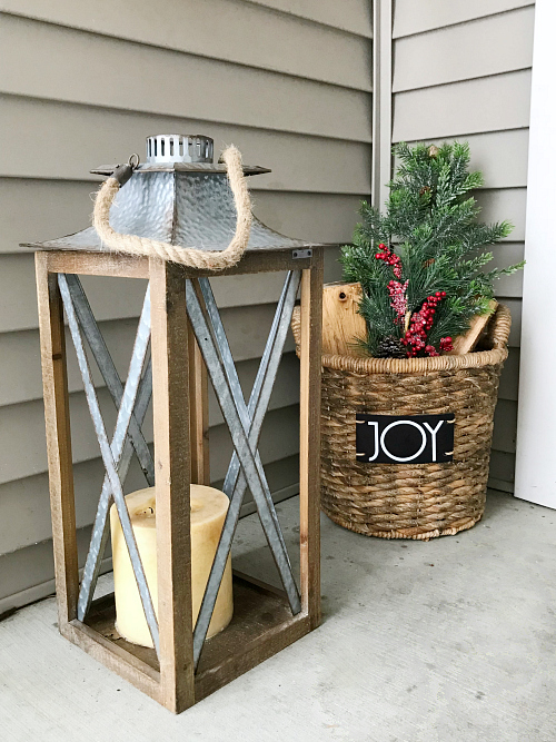 Wood and metal lantern displayed with a round wicker basket filled with firewood and Christmas stems