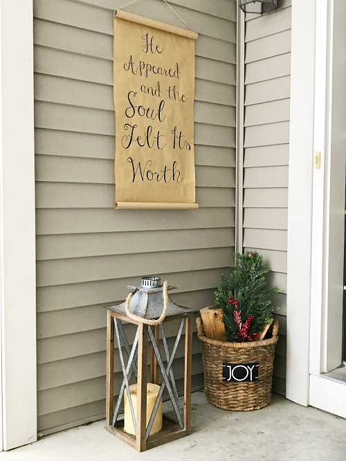 DIY Christmas scroll hung outside on porch wall with basket of wood and lantern below