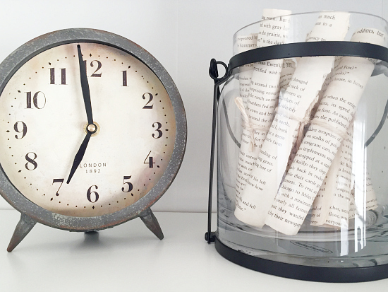 Small clock next to hurricane lantern filled will rolled book pages