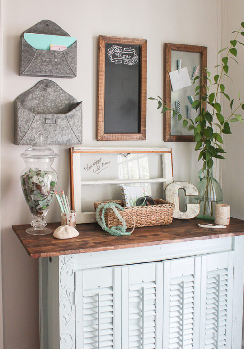 This command center idea come from Sand Dollar Lane and features a wood framed chalkboard, metal-style envelopes, and incorporates the use of a small shuttered table to display an old window and various knick-knacks.