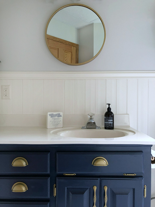 Classic farmhouse bathroom displaying a gold rimmed round mirror above a navy vanity