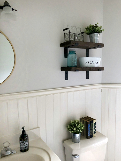 DIY Farmhouse Shelving in a classic farmhouse bathroom. Chunky wood shelves hold various items including a white soap dish and faux plants