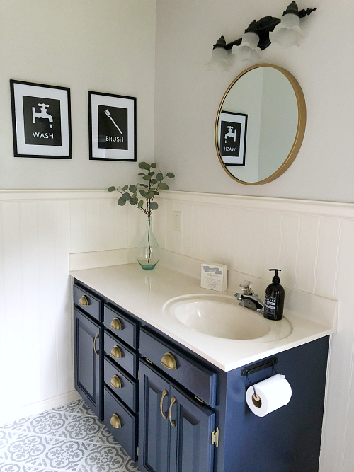 Outdated bathroom gets a classic farmhouse style makeover for less than $100 After image shows DIY wall decor, new mirror, freshly painted navy vanity with brass hardware, and a newly stenciled white and gray patterned floor