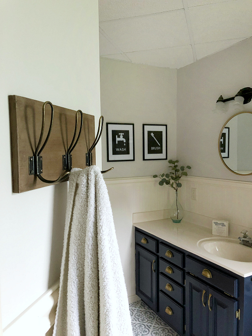 Classic farmhouse bathroom showing DIY towel hooks at the forefront with a navy vanity, stenciled floor, and gold rimmed round mirror in the background