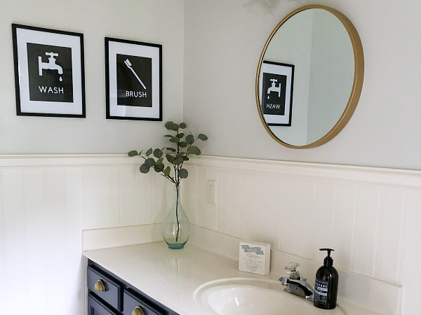 An outdated bathroom gets a classic farmhouse makeover for less than $100. Title image: view of bathroom showing DIY Wall Decor and a round gold mirror above the vanity